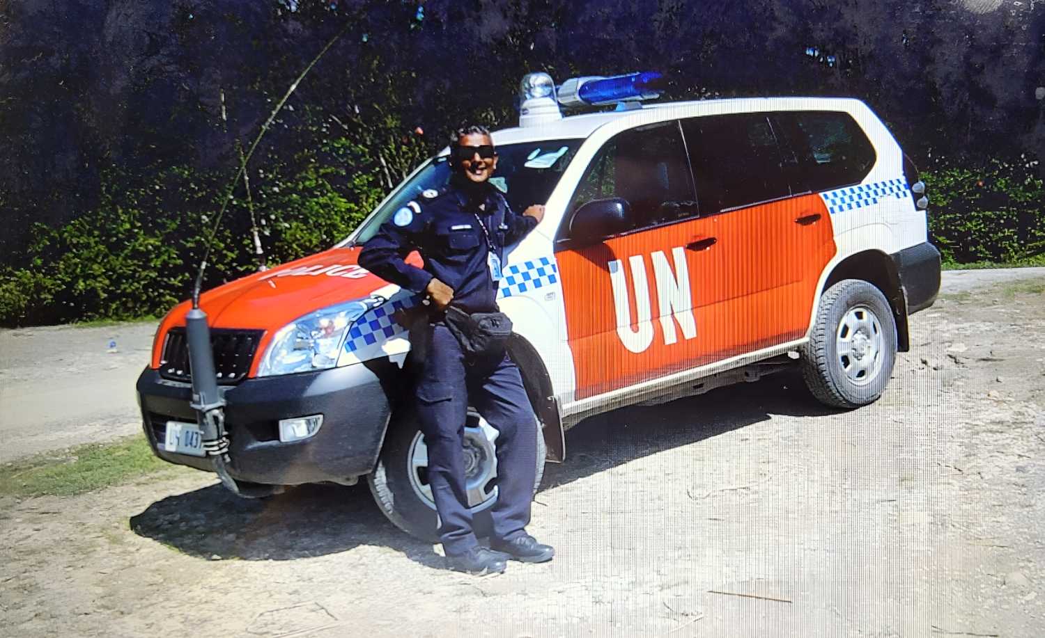DSP Singh standing infront of a UN vehicle. Car is white, hatchback, with a big orange paint on its side only, with the letters "UN" in white decal