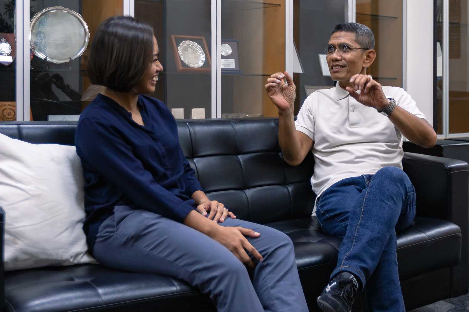 Senior Inspector Julaimi and Sergeant Natassha sitting together on a black sofa while chatting with each other joyfully, both of them have a smile on their face. 