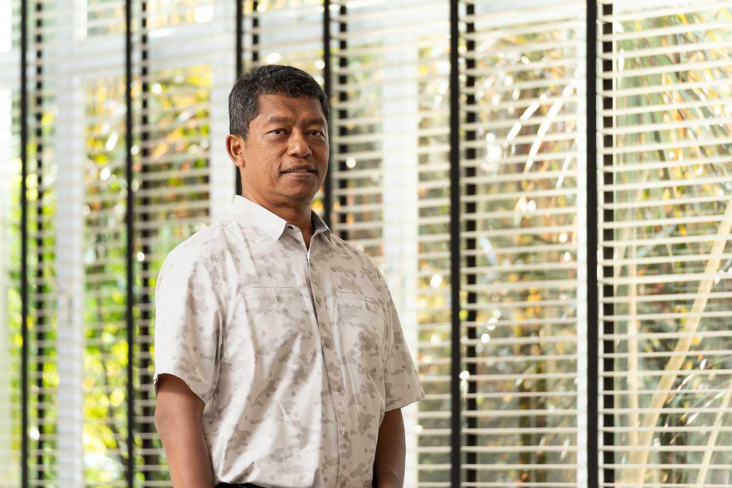 Supt Amir posing for photo beside a window blind. He is a slightly tall man, wearing a white short sleeve shirt, with short black hair with hints of white hair, clean shaven.