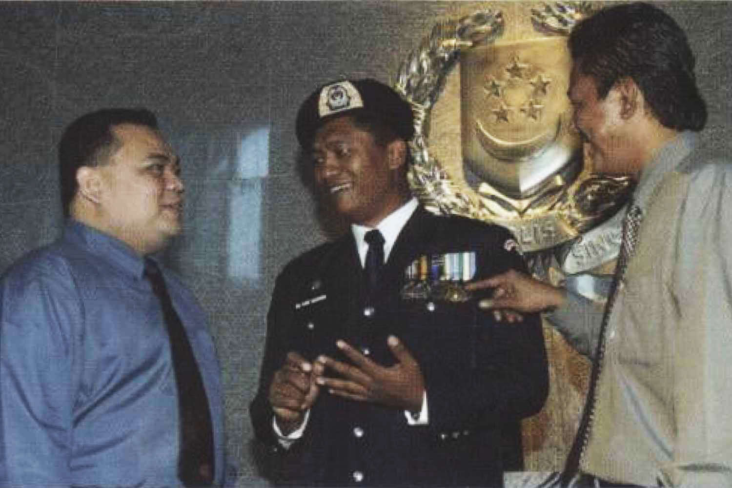 Supt Amir at the New Phoenix Park lobby, infront of a big silver Police Logo, and with two other officers in formal attire. He is wearing a ceremonial attire, with many police ribbons displayed and a beret with the police crest surrounded by yellow.