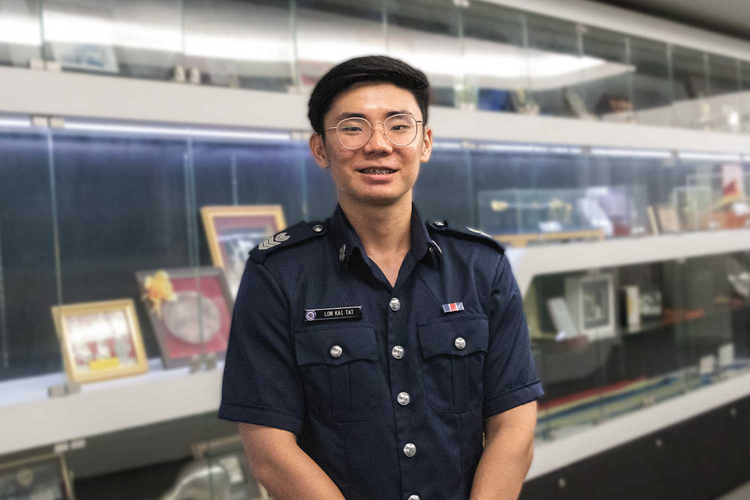 SSSgt Low Kai Tat standing and smiling in front of a wall filled with awards. 