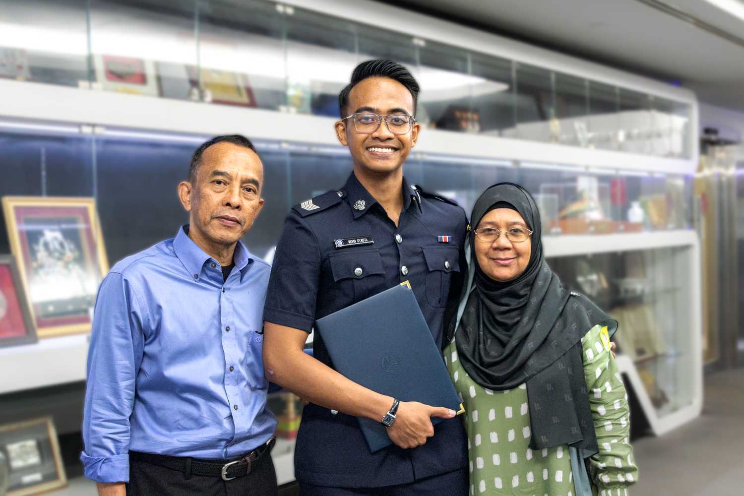 Sgt Muhamad standing and smiling in front of a wall filled with awards, alongside his parents. 