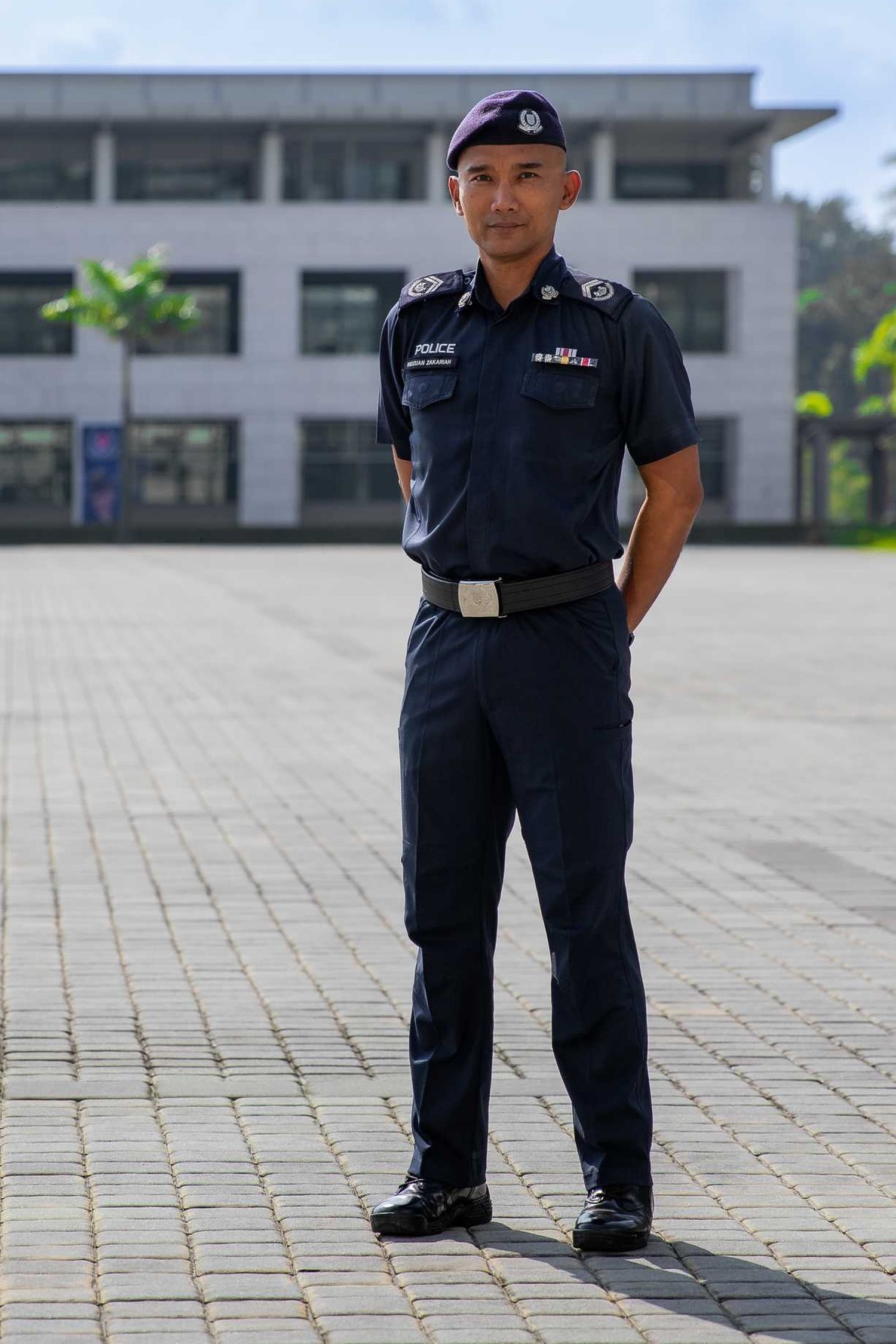 photo of SI Ridzuan, wearing a blue police uniform, on the parade ground with a police building the background. He is slim built, clean shaven.