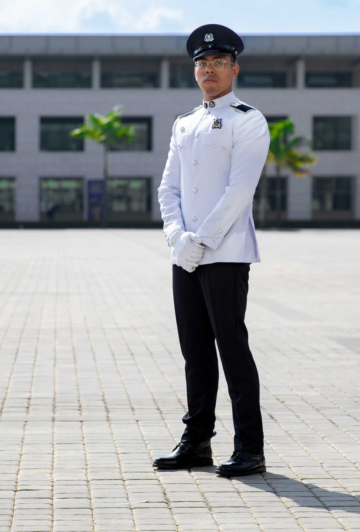 officer is standing in his white uniform, armed clasped infront of him, on the parade ground. He is wearing spectacles and also wearing a peak cap