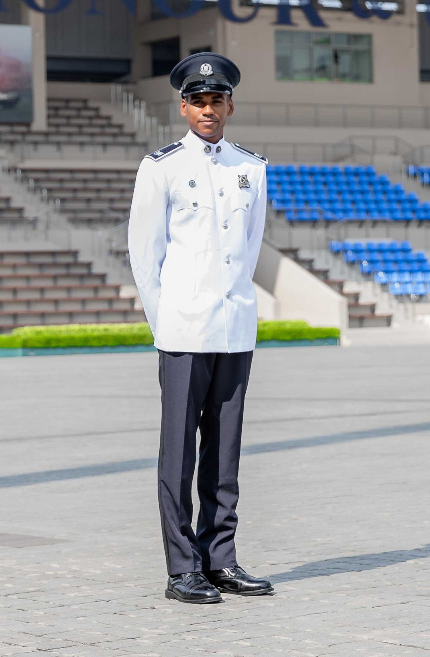 officer is standing on the parade ground with the stands behind him. He is wearing a white ceremonial attire with a dark blue peak cap, with his arms firmly to the back. 