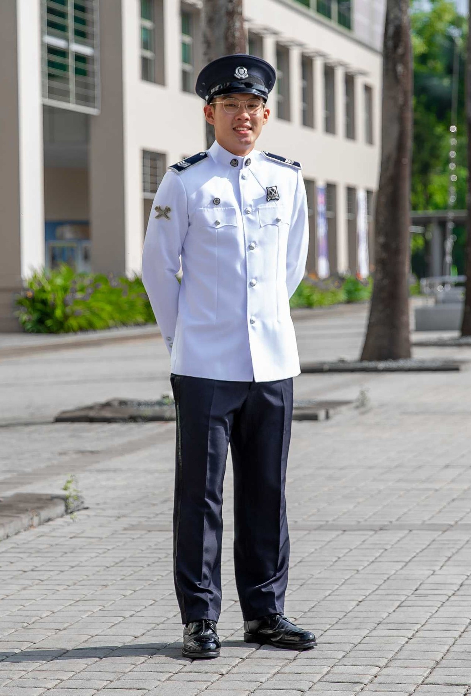 officer in white uniform and dark blue pants, standing with this arms clasped behind. He is on the parade ground, with some very tall trees in his background