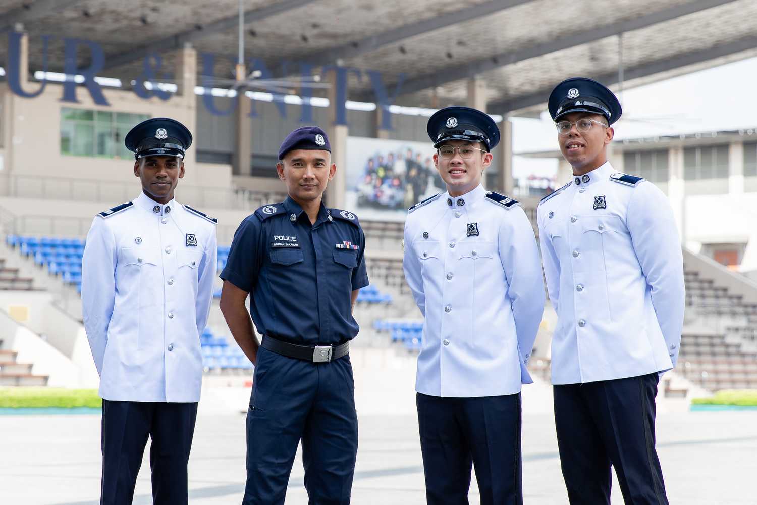 Four officers standing on a parade ground, where the left most extreme is wearing a ceremonial white attire, 2nd is wearing a blue police uniform, and the rest are wearing a ceremonial attire