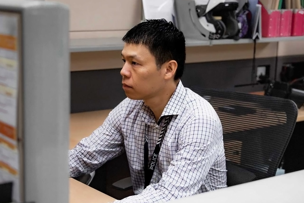A/Insp Adrian Lim sitting in office wear at his desk, wearing his SPF lanyard. His hands are out of reach and covered by a wall, presumably typing on a computer.