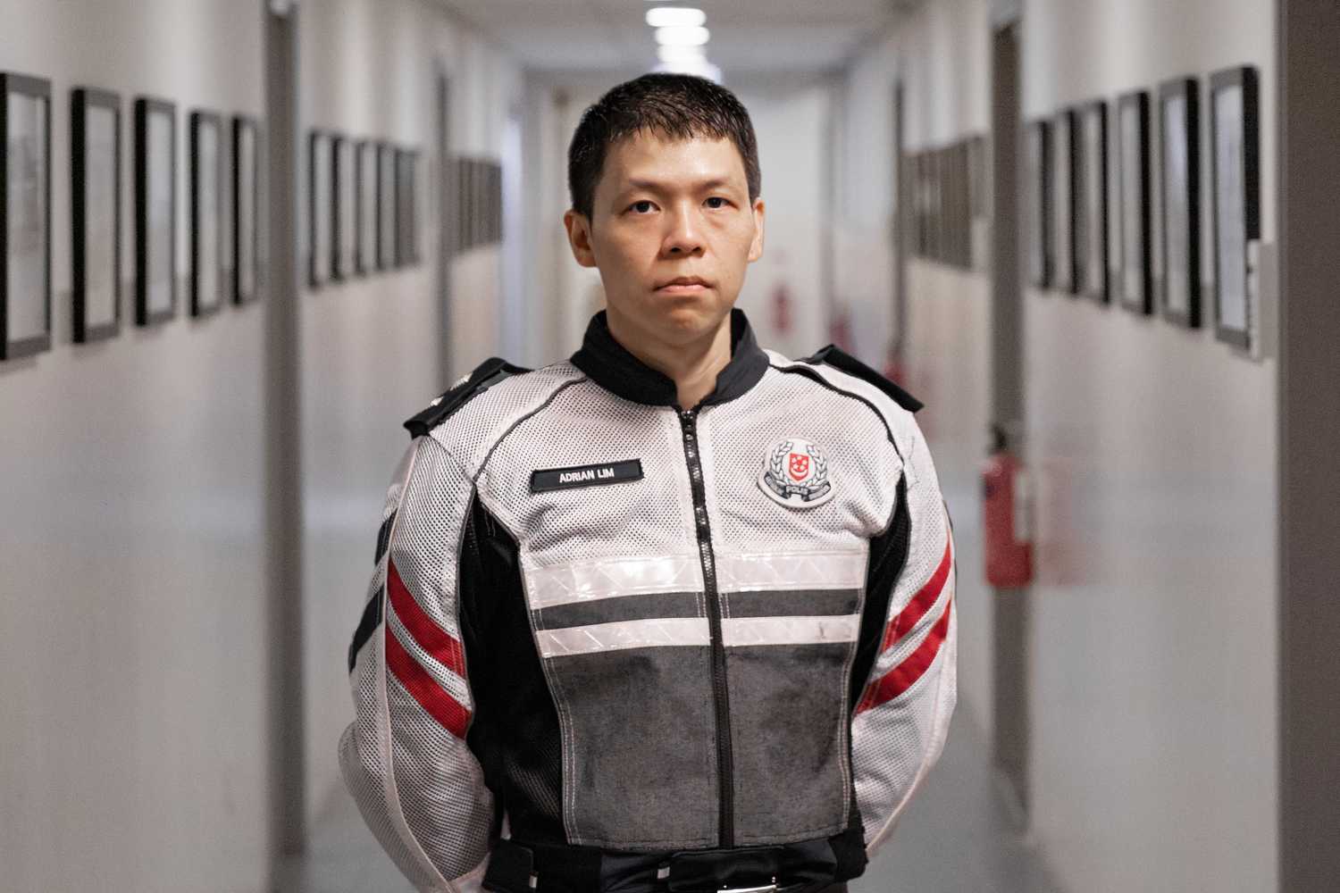 A/Insp Adrian Lim standing in front of a corridor filled with photo frames, with his hands behind his back, looking straight at the camera in a formal shot.