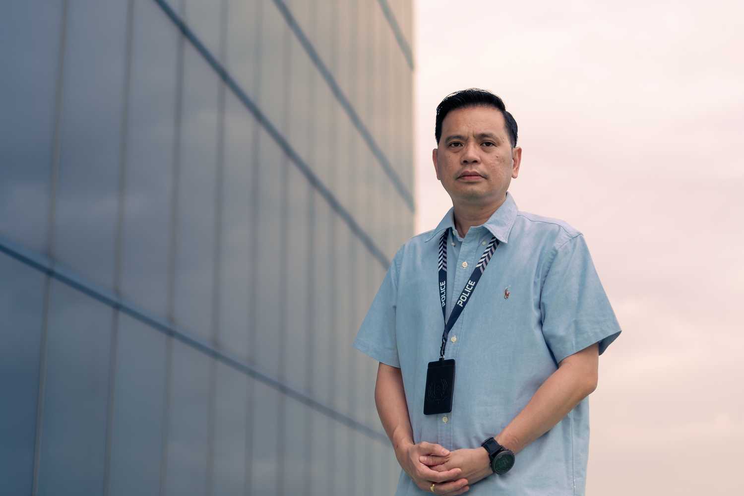 photo of officer wearing blue shirt with arms infront, standing infront of a building behind him. The sky is setting in the background. 