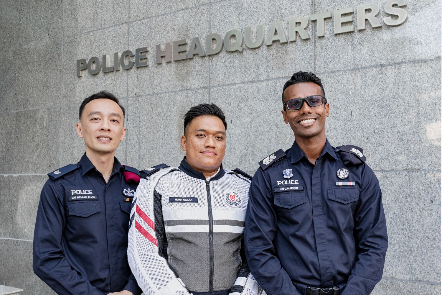 ASP Alvin Lai, Sgt Muhammad Adzlan Bin Hussein and SSSgt Mohamed Hafiz Maideen posing side by side, smiling and looking at the camera underneath a sign that says 'Police Headquarters'. They are in uniform