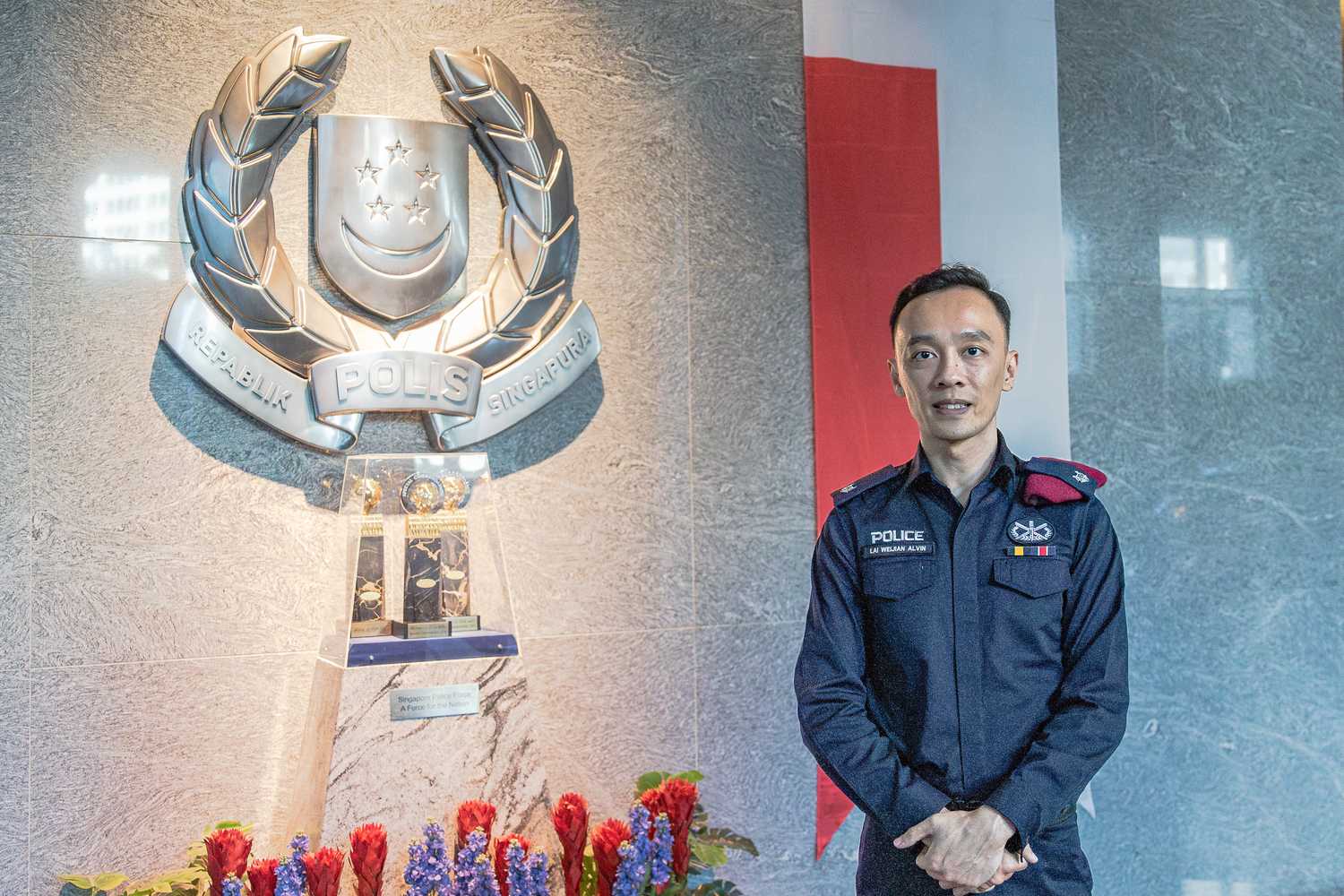 ASP Lai with his hands folded in front of him, standing beside a Singapore Police Force crest and looking proudly at the camera