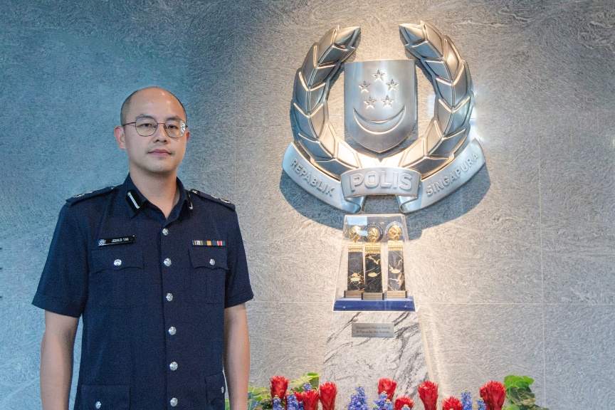 Assistant Commissioner of Police (AC) Jerald Tan, Commander of Jurong Police Division, standing and looking proudly at the camera in front of a large Singapore Police Force crest mounted on the wall.