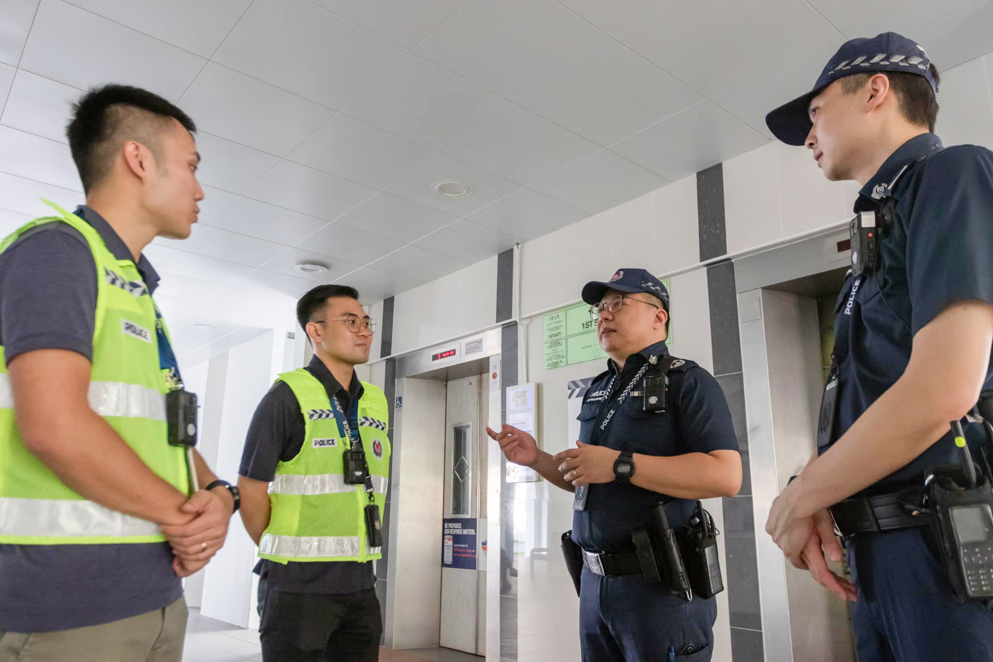 Four Singapore Police Force officers are conversing with each other in the lift lobby of a Housing Development Board flat. Two are in uniform, while the other two are wearing police vests and police lanyards over plainclothes.