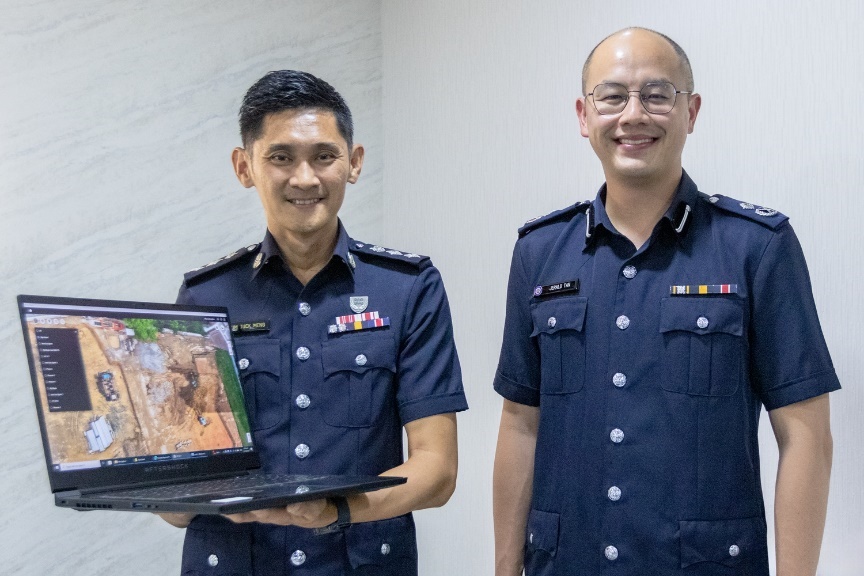 Superintendent of Police (Supt) Sum Tuck Meng, Head of UAV Operations and Training at the Home Team UAV Unit is standing beside Assistant Commissioner of Police (AC) Jerald Tan, Commander of Jurong Police Division. Superintendent of Police (Supt) Sum Tuck Meng is holding a laptop displaying the photo of the drone-created 2D map overlay, both officers are looking proudly at the camera.
