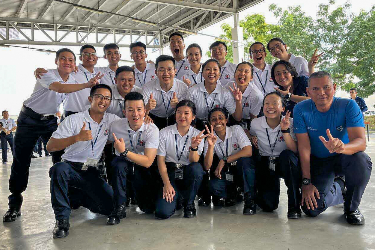 A squad photo of ASP Shermaine Ang and ASP Cammy Carin Chua during their training days. They and their squadmates are standing or squatting in three rows and pulling funny faces, looking happy.