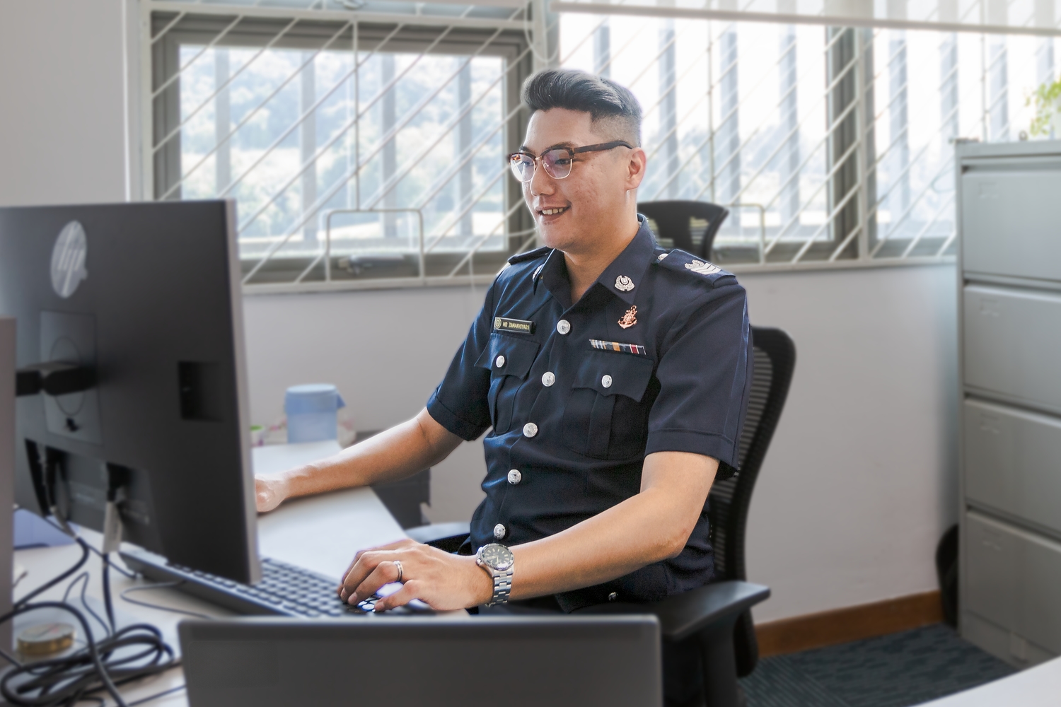 Senior Staff Sergeant (SSSgt) Muhammad Zamakhsyarii is sitting at his desk, looking at his computer with his hands extended on the keyboard, as if he is about to type something.