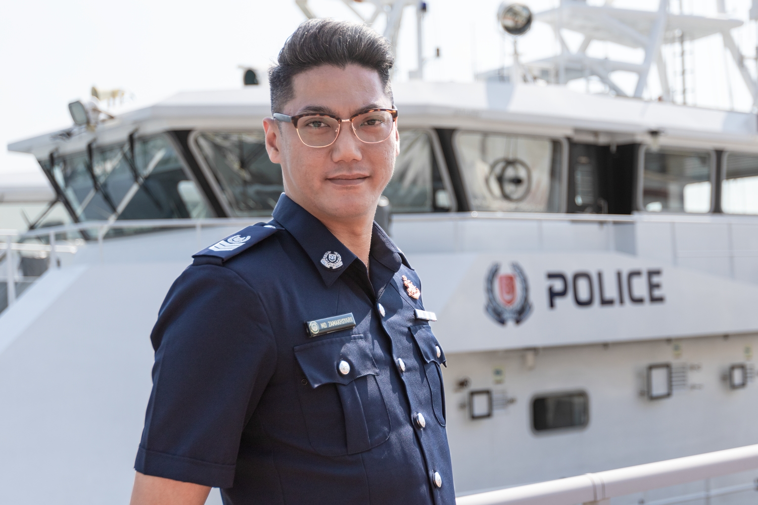 Senior Staff Sergeant (SSSgt) Muhammad Zamakhsyarii in a profile shot, looking proudly at the camera. In the background, there is a police boat with the Singapore Police Force logo on it.