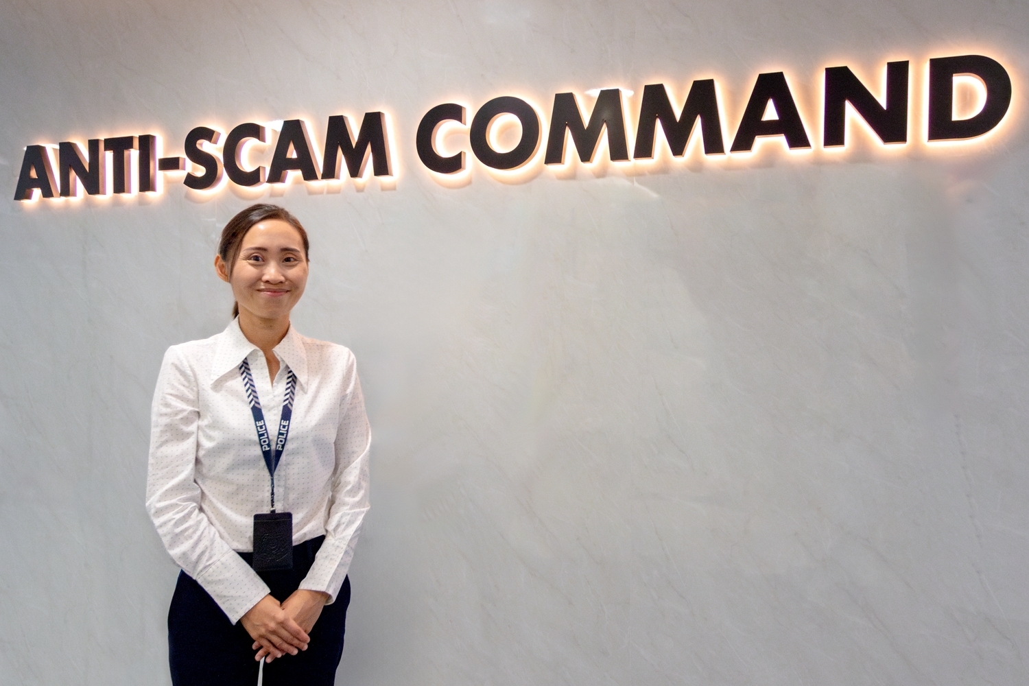 A profile shot of DSP Clara Wong, standing below a sign on a wall that reads 'Anti-Scam Command'. She is smiling at the camera.