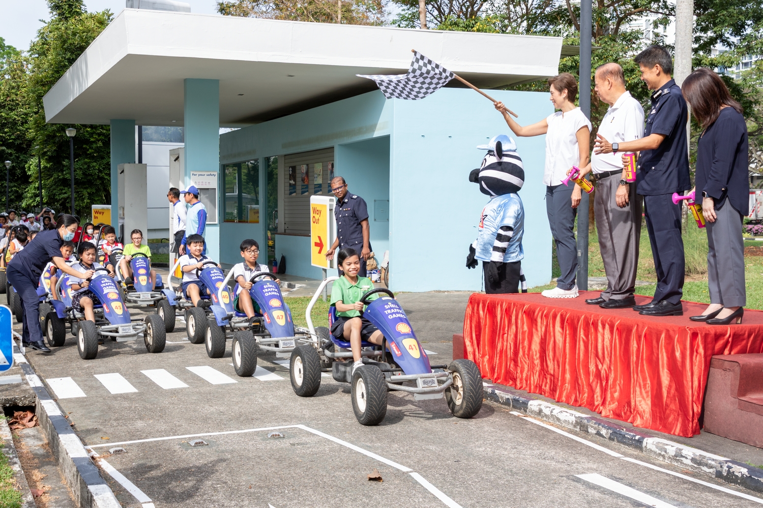 Happy primary school children are sitting in go-karts and pedalling off after the start of the race has been announced through air horns and a flag off. Standing on podium is Mrs Josephine Teo, Minister for Communications and Information & Second Minister for Home Affairs, holding a flag to signal the start of the race. Next to her are Mr Bernard Tay, Chairman of the SRSC, Senior Assistant Commissioner of Police Daniel Tan, Commander of Traffic Police, and Mdm Susan Wang, Superintendent East 2, Schools Division, MOE, who are holding airhorns to signal the start of the traffic games.