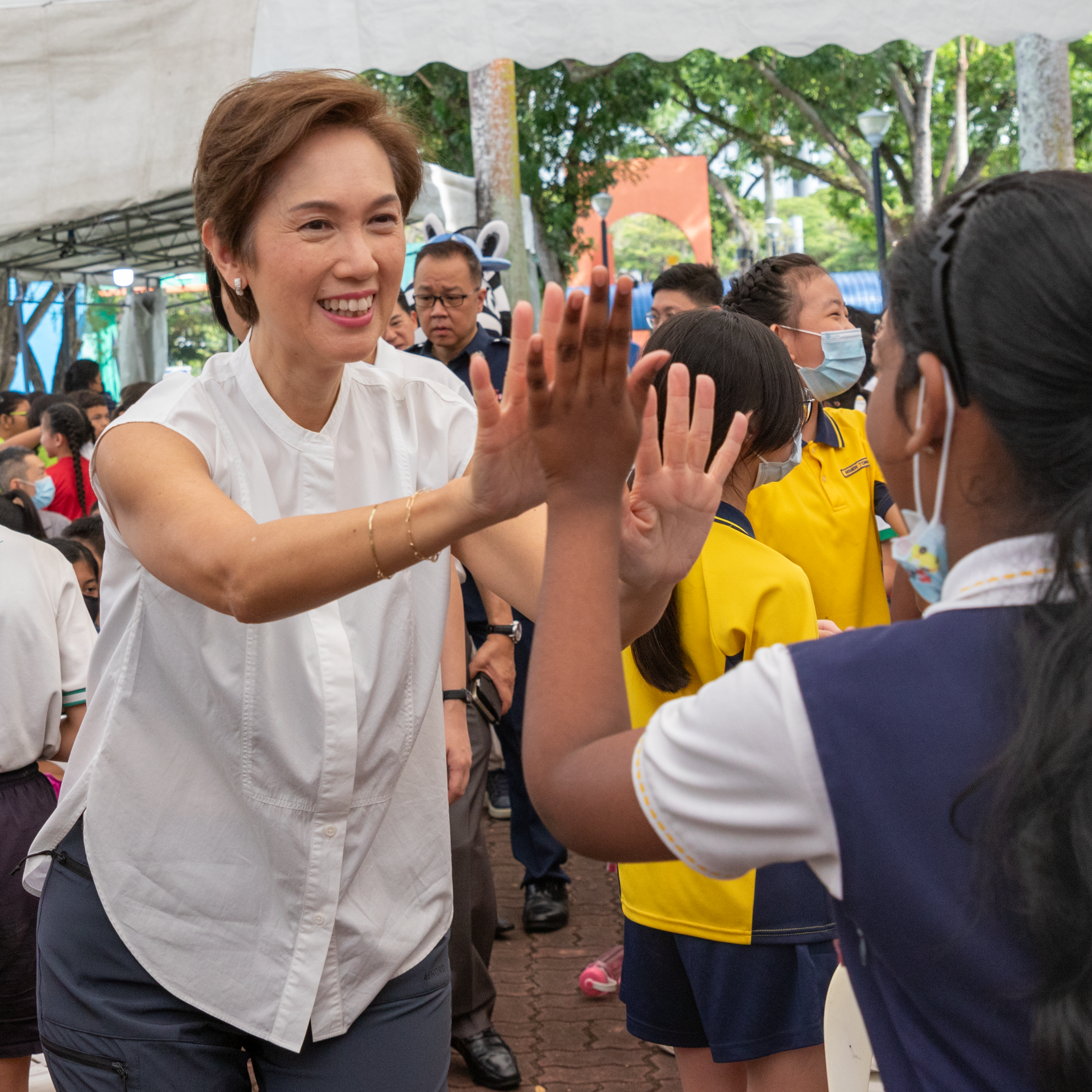 Mrs Josephine Teo, Minister for Communications and Information & Second Minister for Home Affairs, is happily greeting a primary school girl by extending her hands out to her. The girl is also connecting her palms with Mrs Josephine Teo's palms in a high-five gesture.
