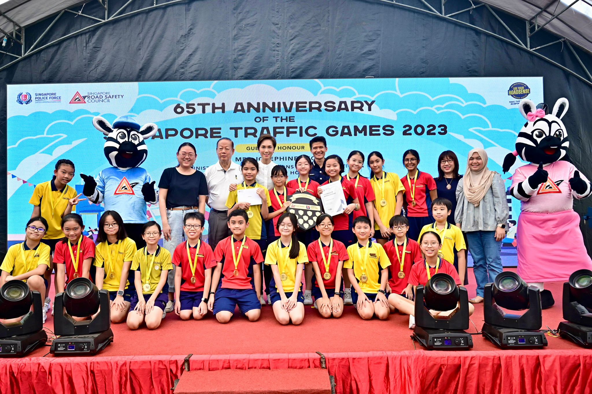 Mrs Josephine Teo, Senior Assistant Commissioner of Police Daniel Tan, Mr Bernard Tay, and Mdm Susan Wang standing on stage with the Champion of the Singapore Traffic Games Challenge Trophy 2023, Elias Park Primary School. The students are donning gold medals and smiling proudly alongside the zebra mascots.