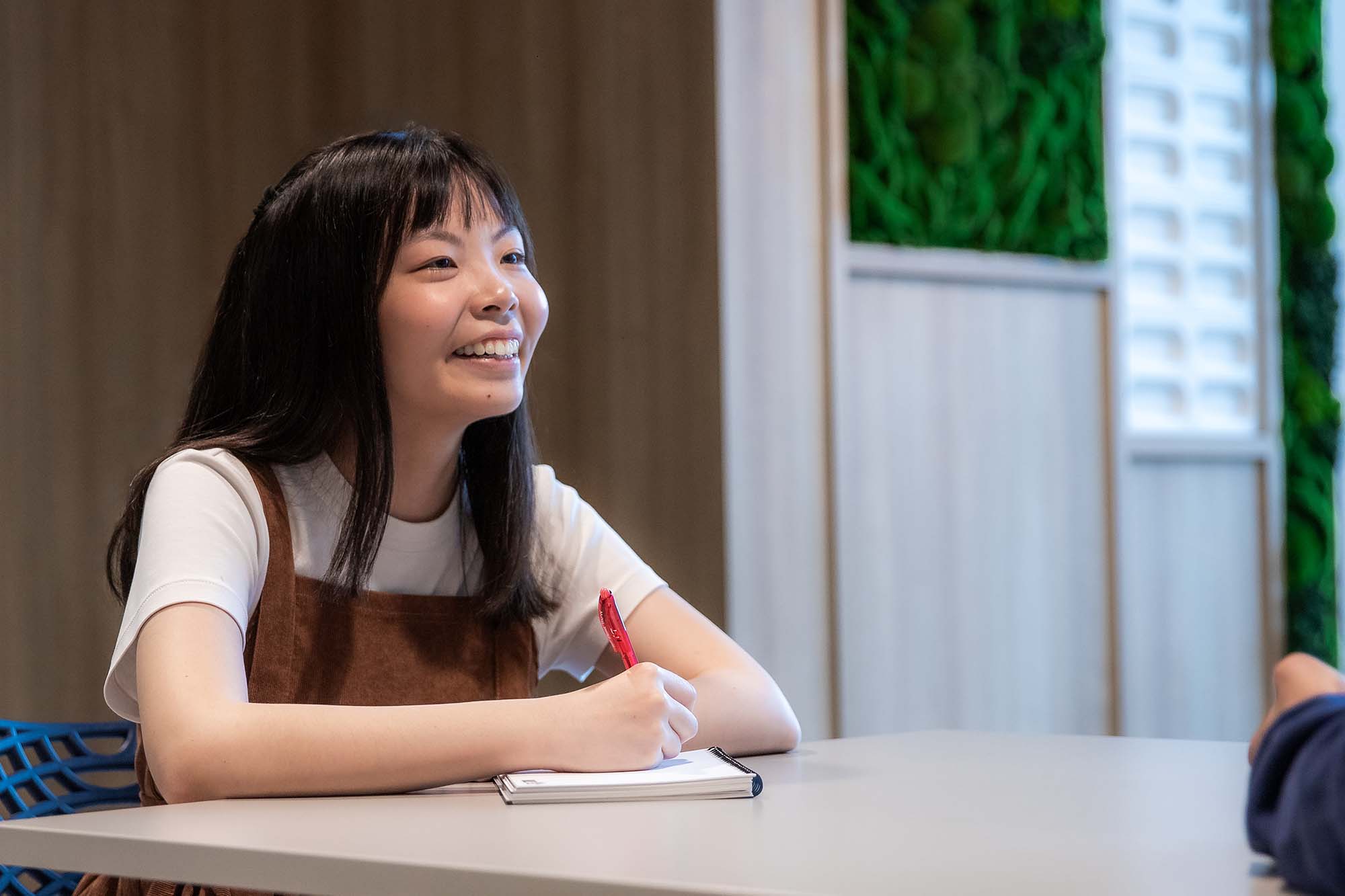 A portrait shot of Amanda Wong shows her holding a pen and jotting down notes on her notepad. She is also intently listening to an interviewee.