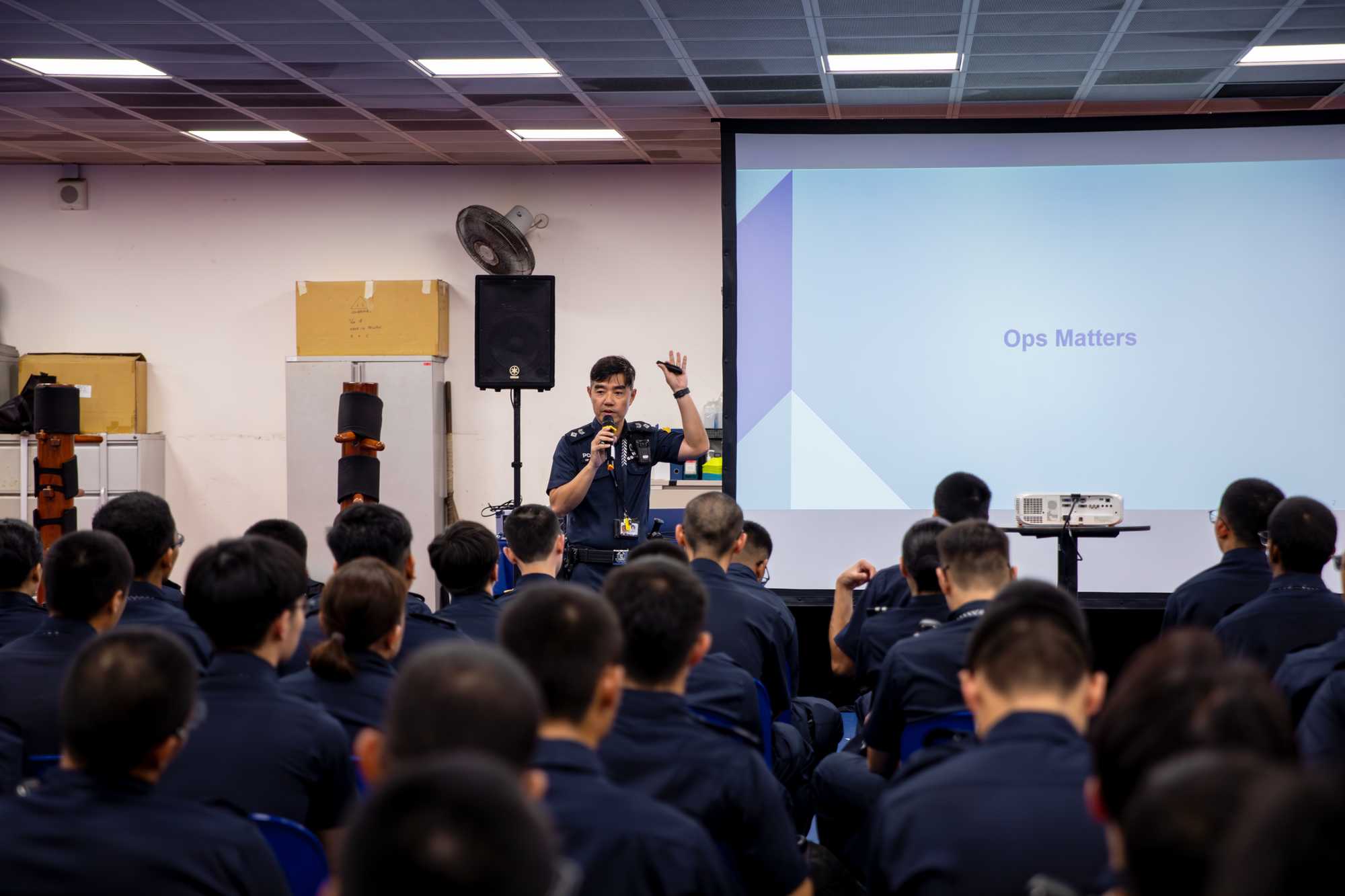 photo is from the back of a big room, where you can see the many heads of officers looking towards one officer who is standing and conducting infront of a projector slide which shows "Ops Matters"