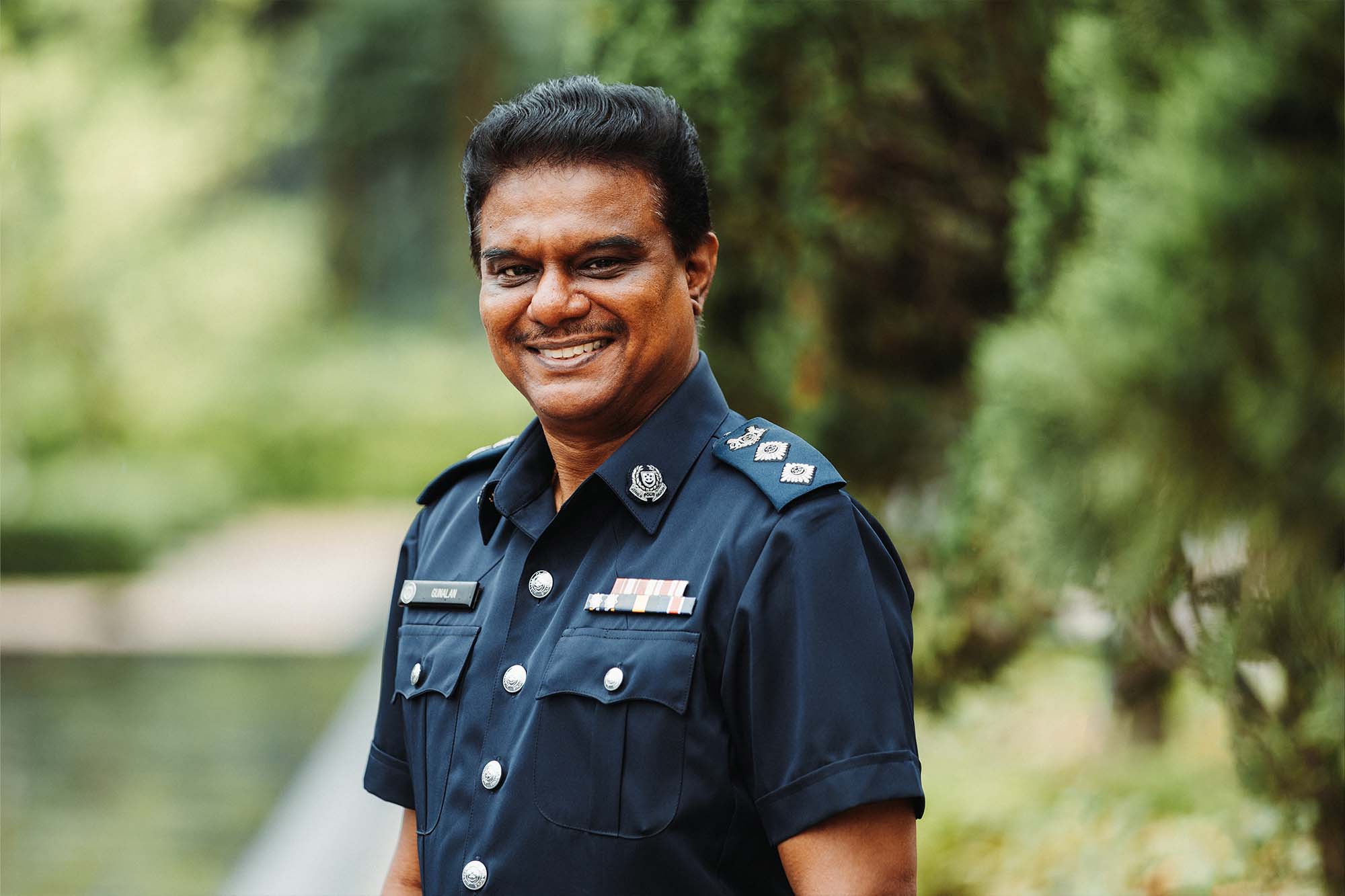 supt gunalan standing alone in the photo smiling with greenery at the background out of focus
