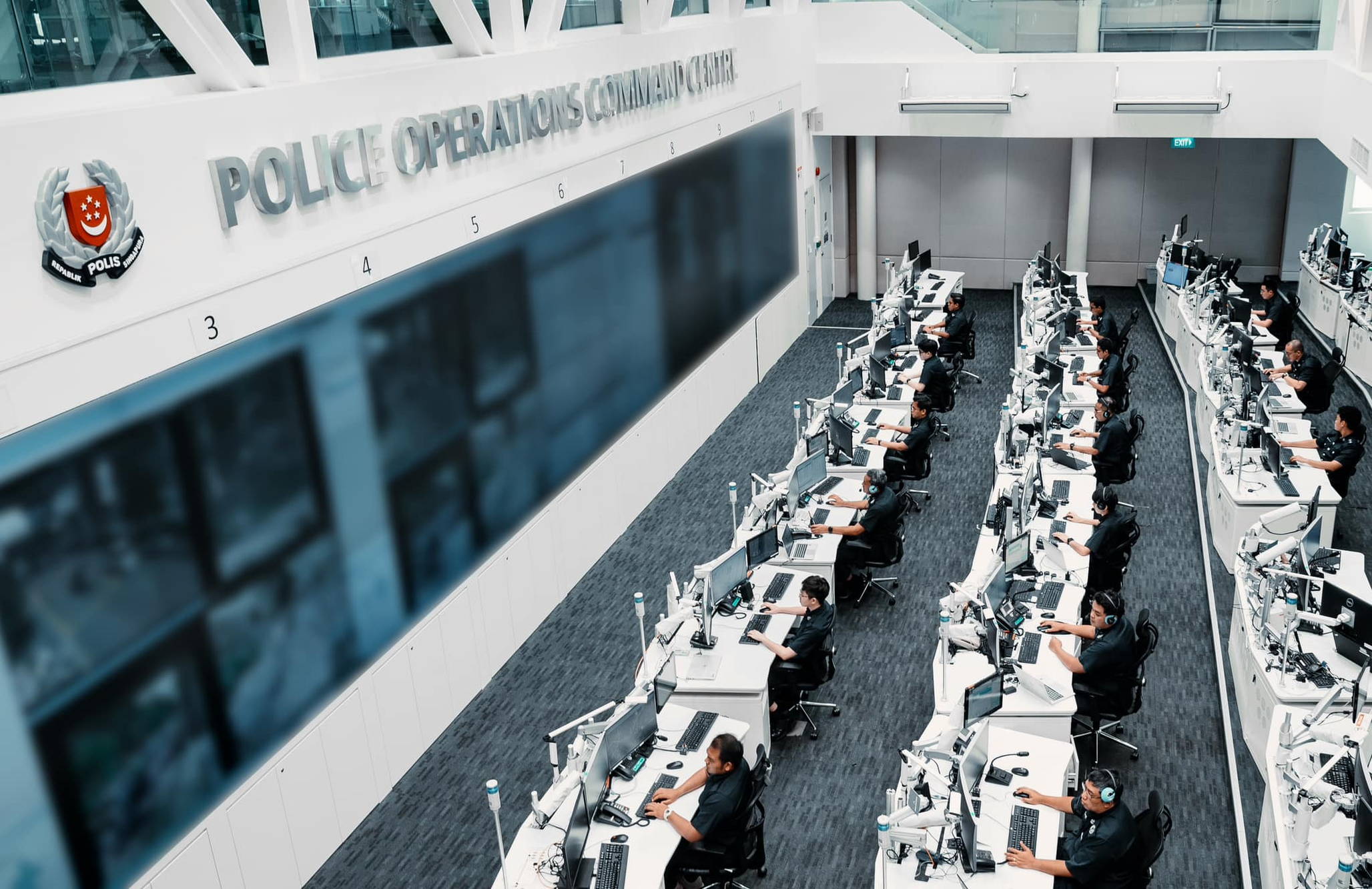 a top down photo of the pocc where officers are seen sitting at desks and answering calls. they are facing left and infront of them is a huge screen that is blurred out