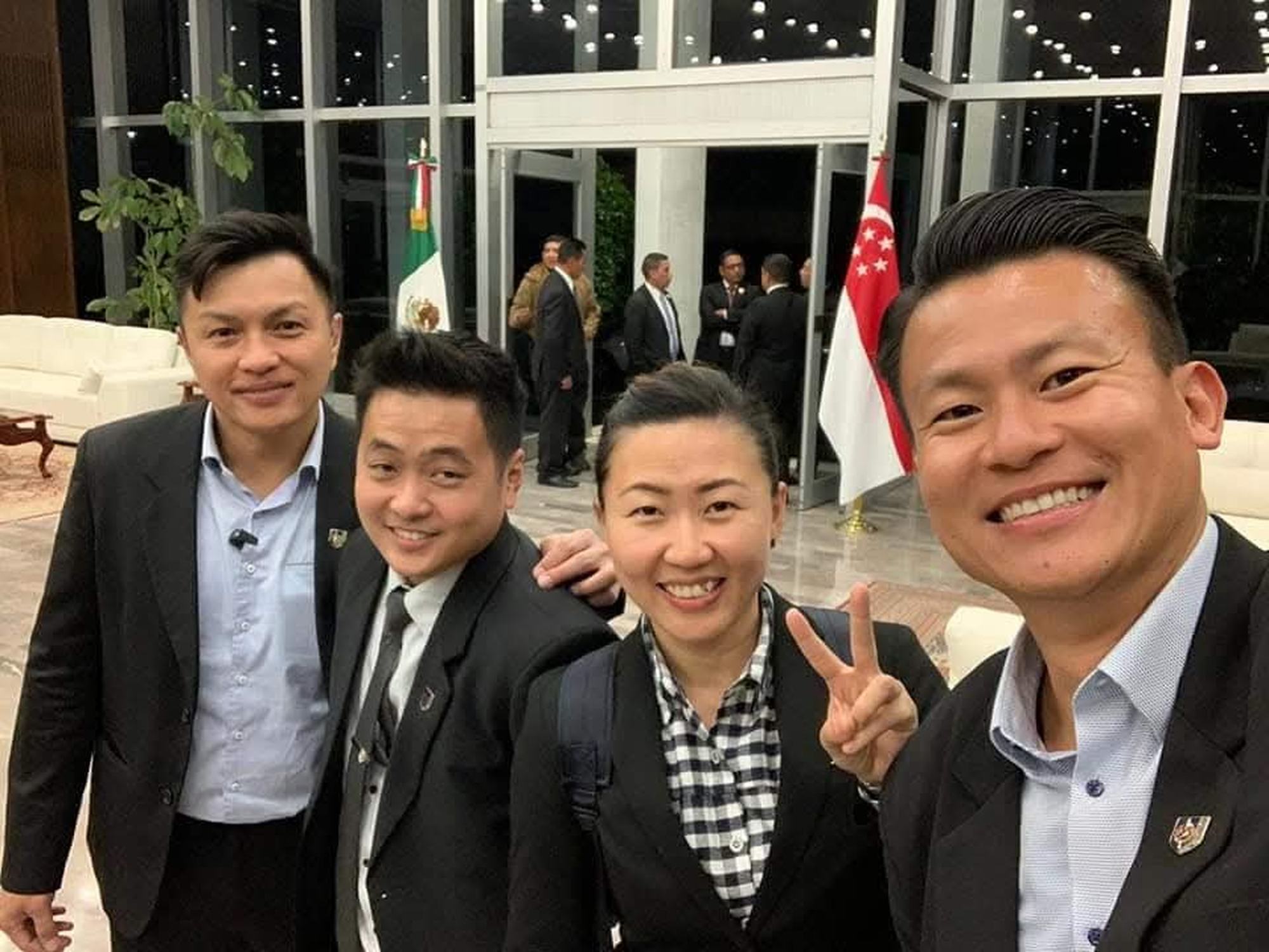 four officers in black blazers and formal shirts in an airport setting, with two flags, of singapore and mexico behind respectively.