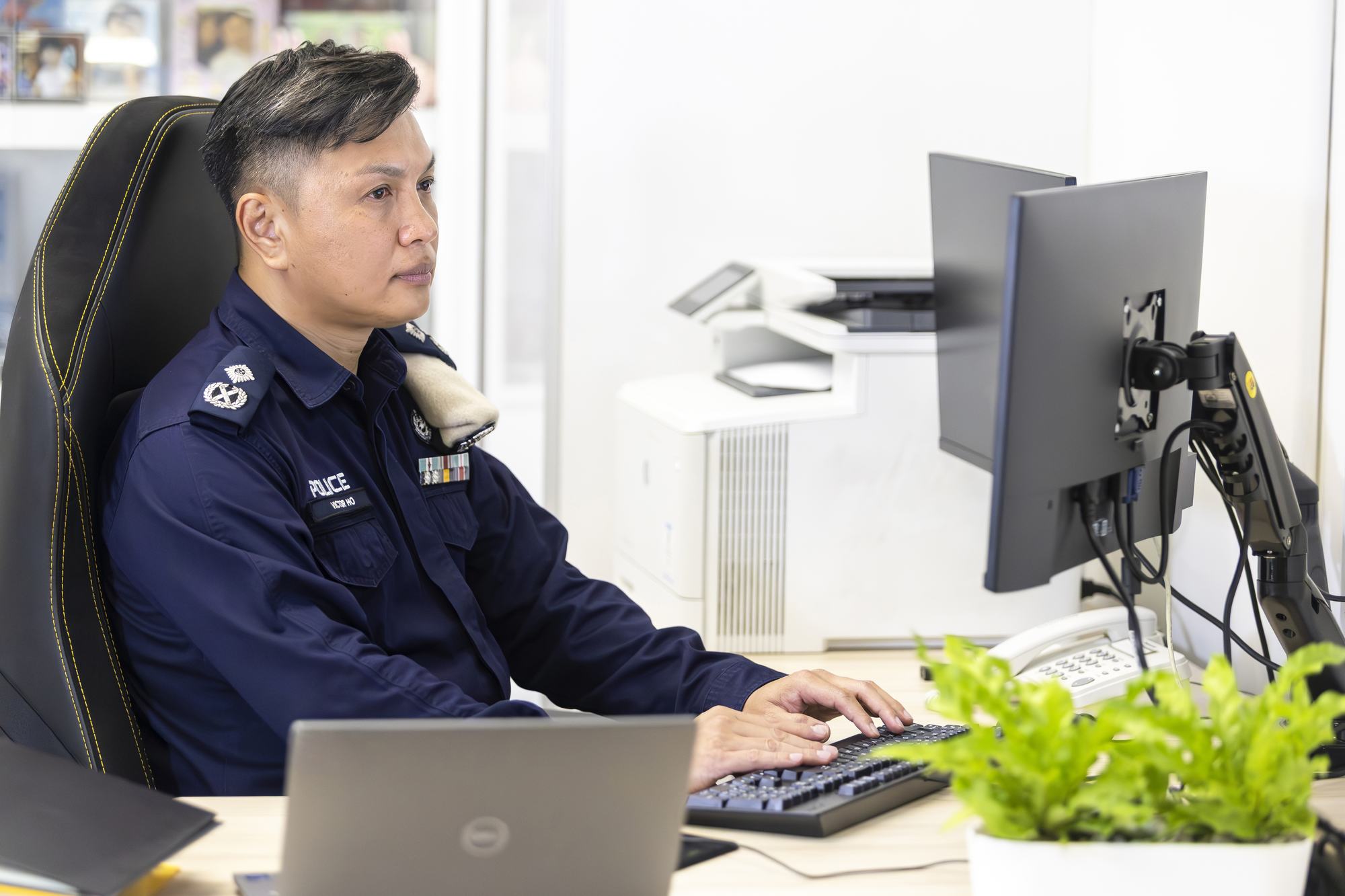 ac victor ho by his desk, facing right, using his computer and typing on the keyboard. His office is well lit and he has a small plant near his monitor