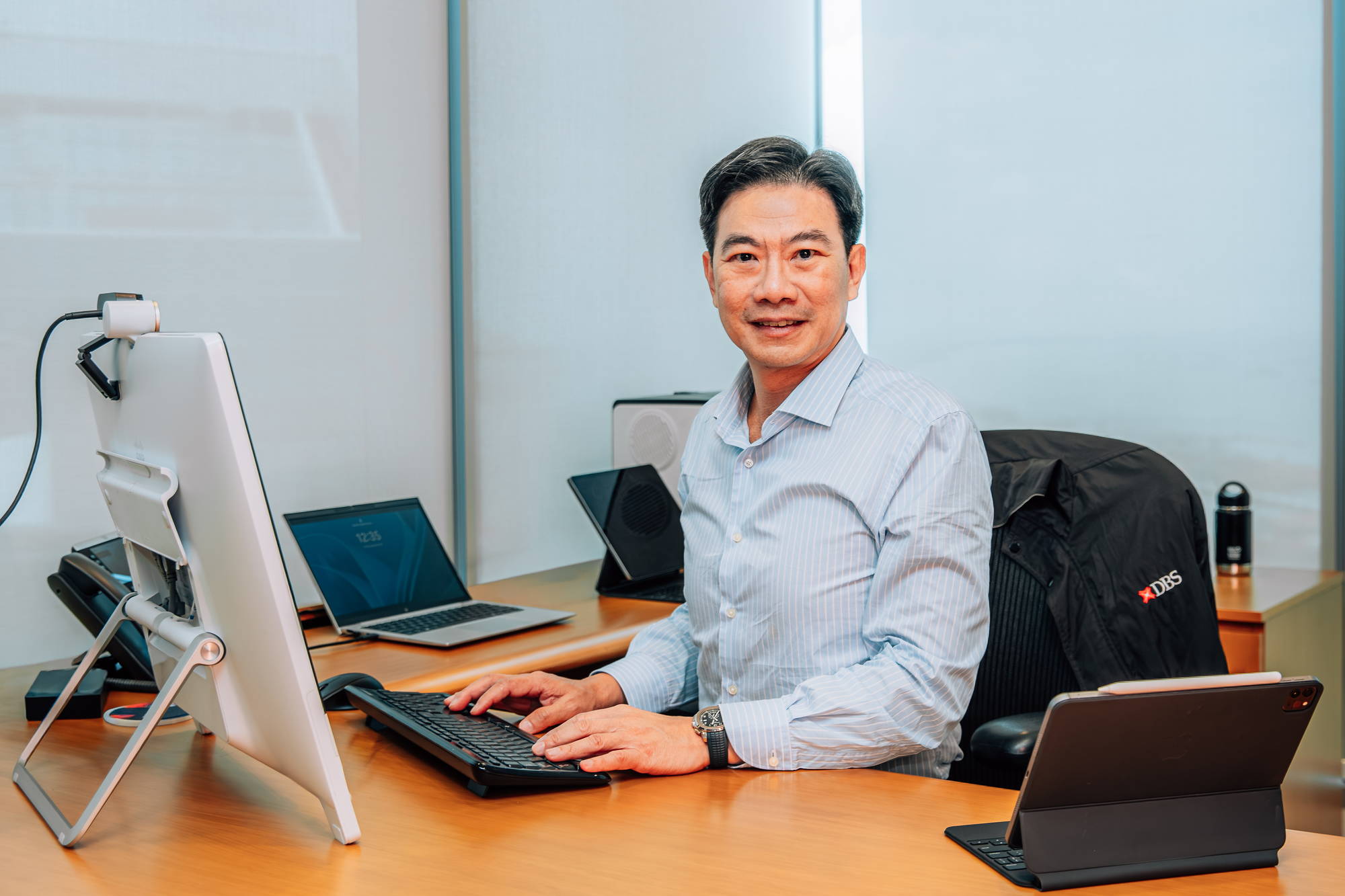 mr derrick goh wearing a white top behind his desk; fingers on the keyboard; smiling for the camera