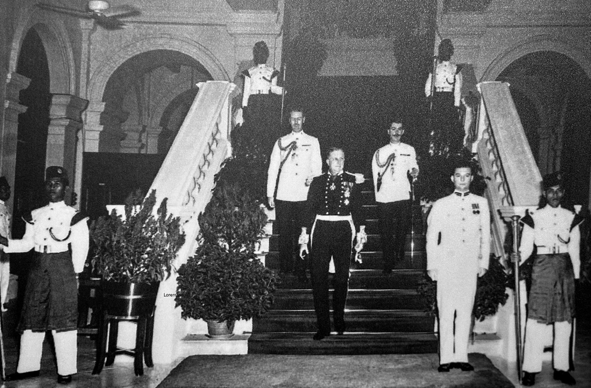 an old photo of a colonial building with a wide flight of stairs. On it the governor, in ceremonial clothing, is walking down, surrounded by officials. Photo is monochromed.