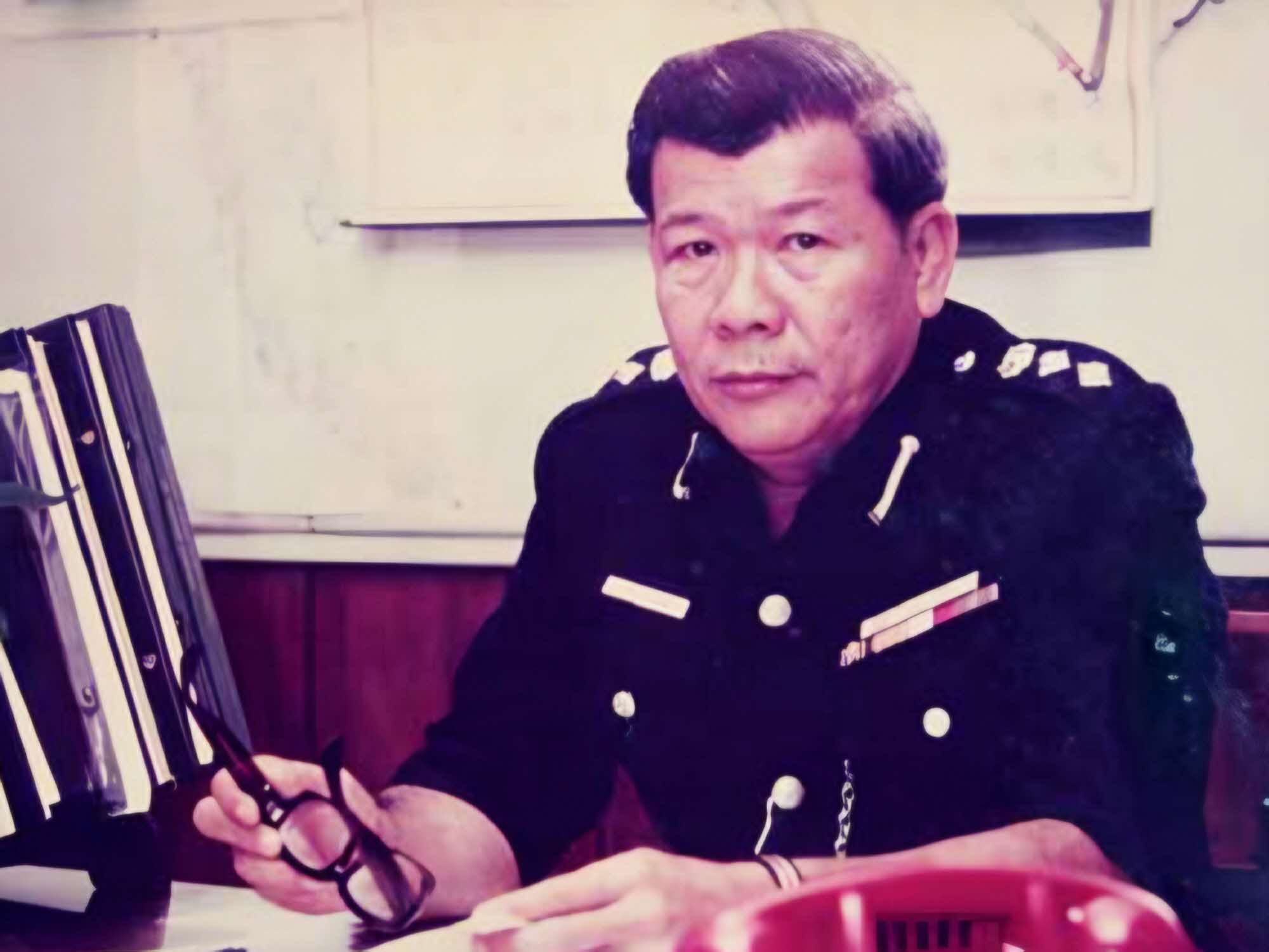 officer in uniform, sitting behind a desk, holding onto his spectacles. On the desk are files and a red phone
