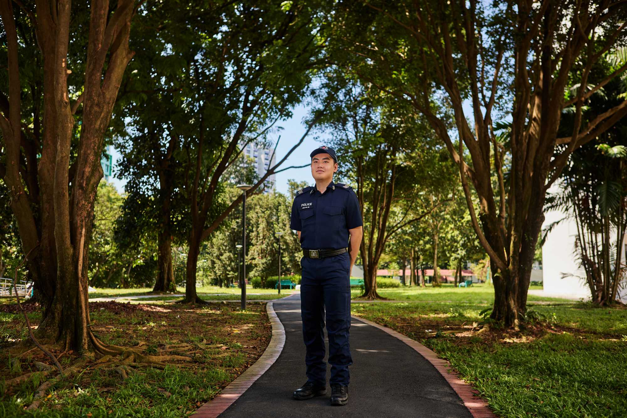Alastor in uniform standing in the middle of a park posing for shoot purpose. He is smiling and has his hands behind his back.