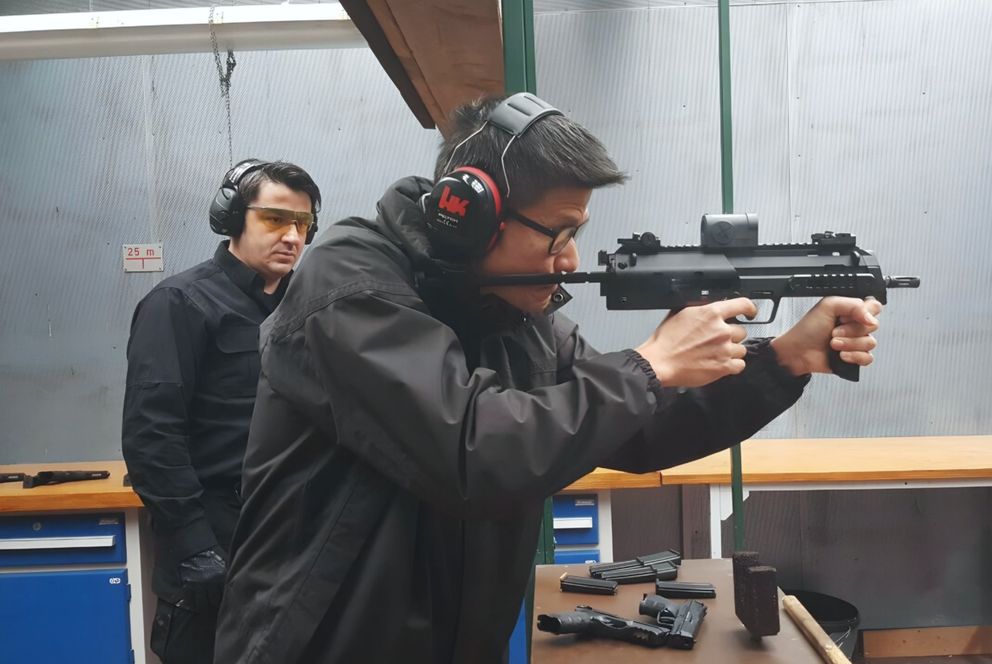 SAC Daniel Tan in a windproof jacket holding an assault rifle at firing at the right side. He is in a firing range with a Caucasian instructor beside him