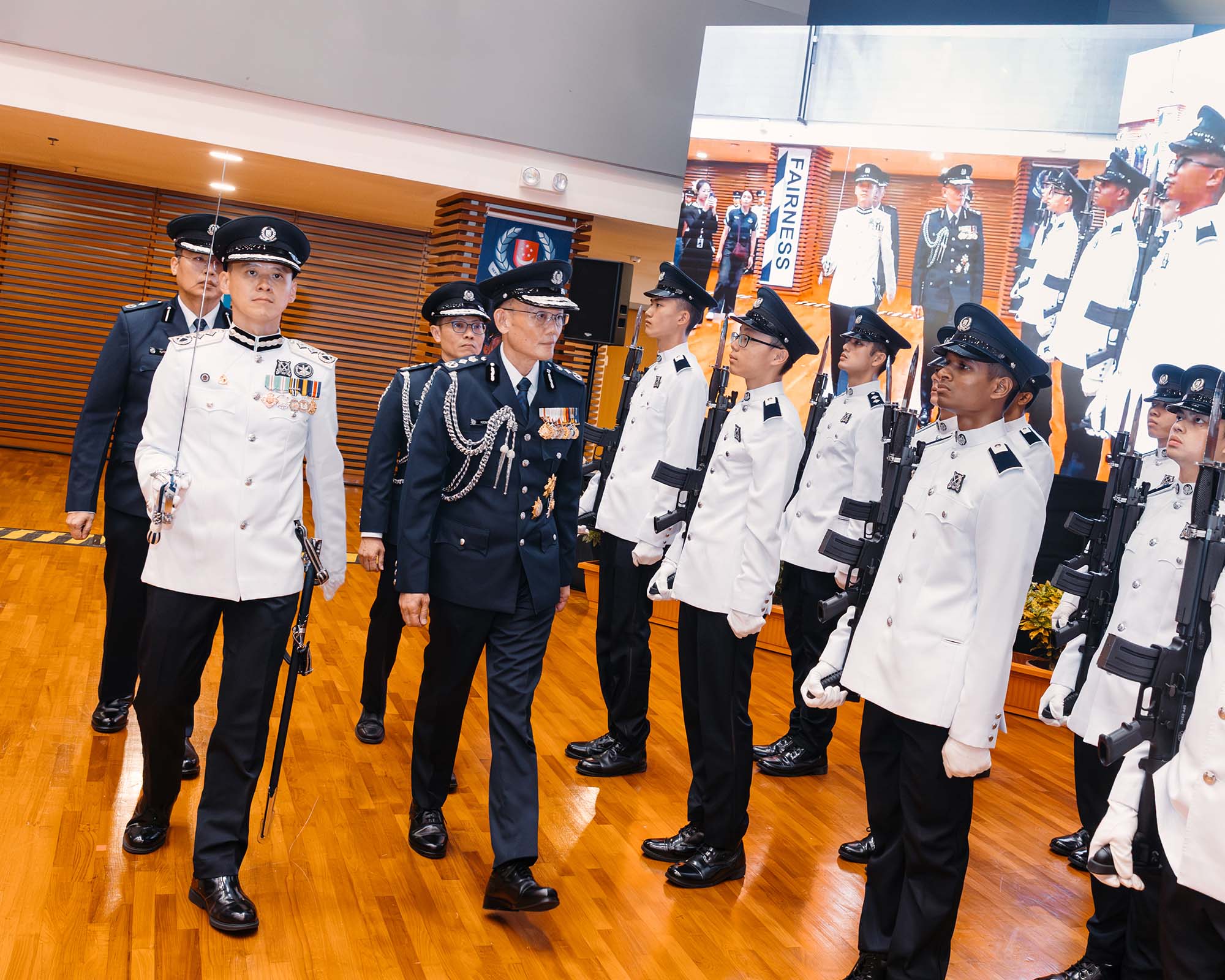 CP hoong Wee teck walking past officers in ceremonial white attire, standing in attention. They are in an enclosed hall, with wooden flooring