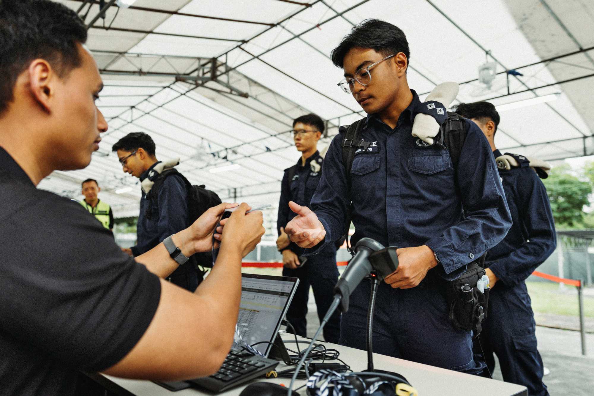 a procom officer in uniform standing infront of a booth and registering his attendance. they are under a white tentage