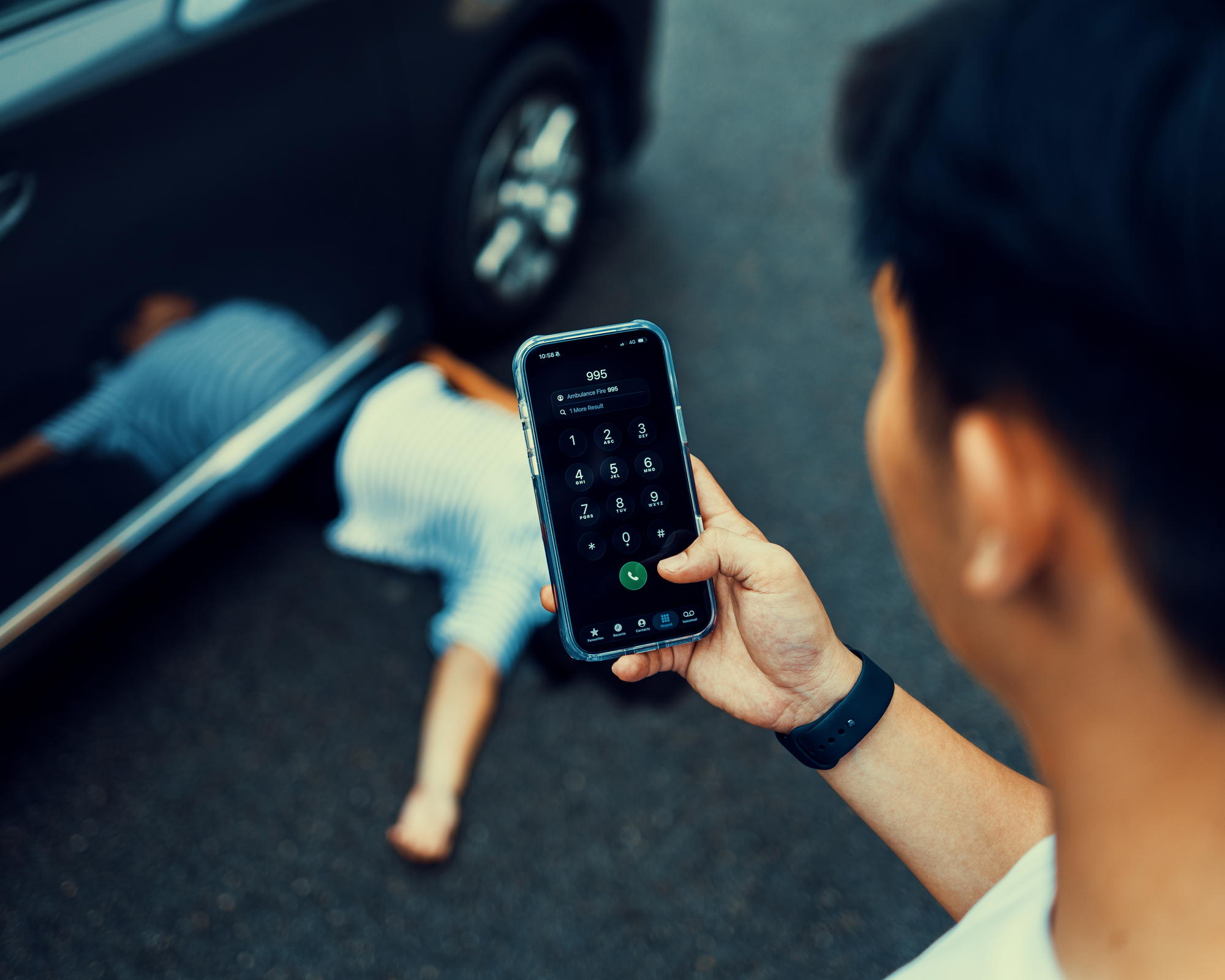 a photo showing a lady lying on the road under a car and a man using a phone to call 995