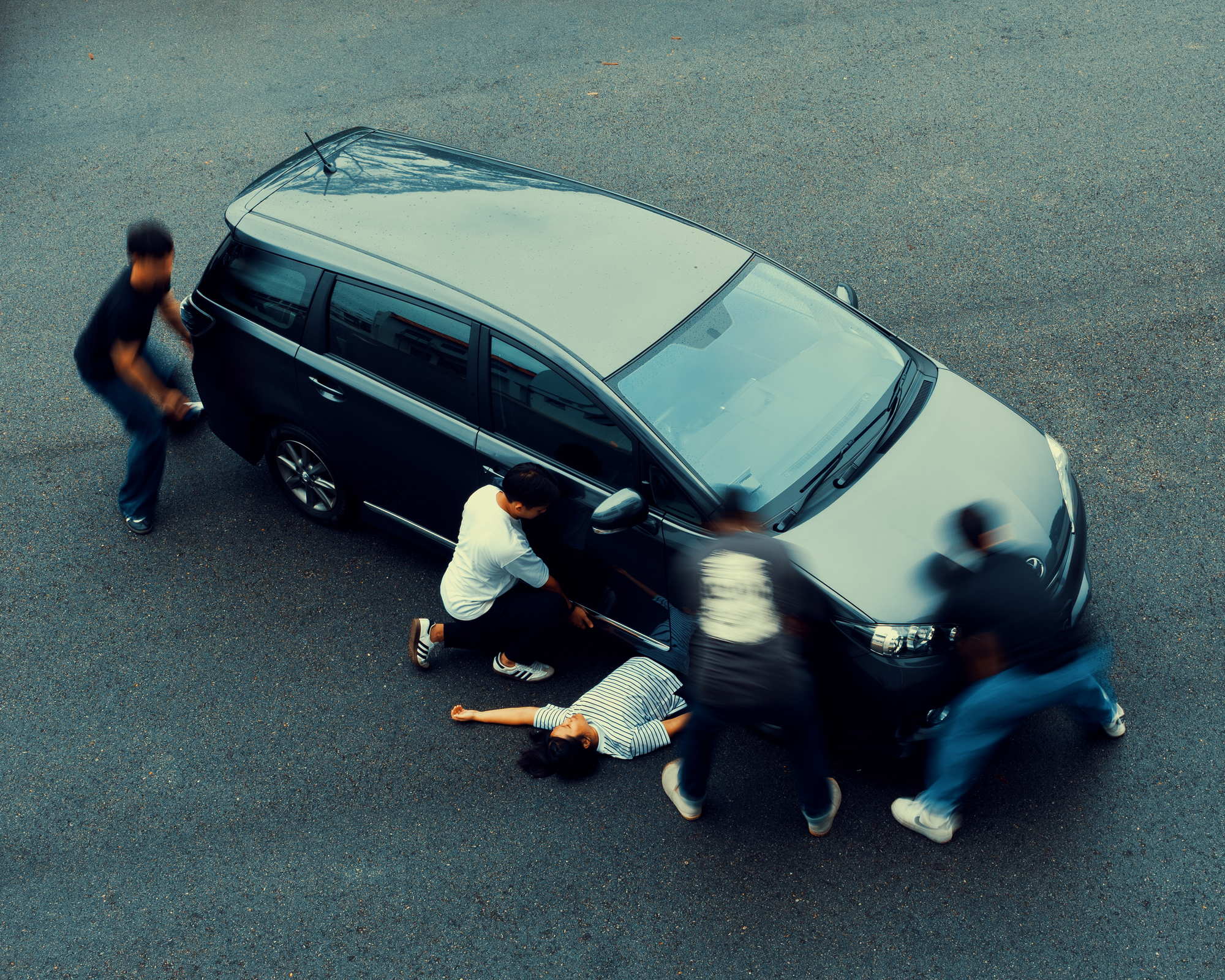 there are fast moving human figures around a black car shown to be finding positions to lift the car. A lady in white is lying on the road under the car