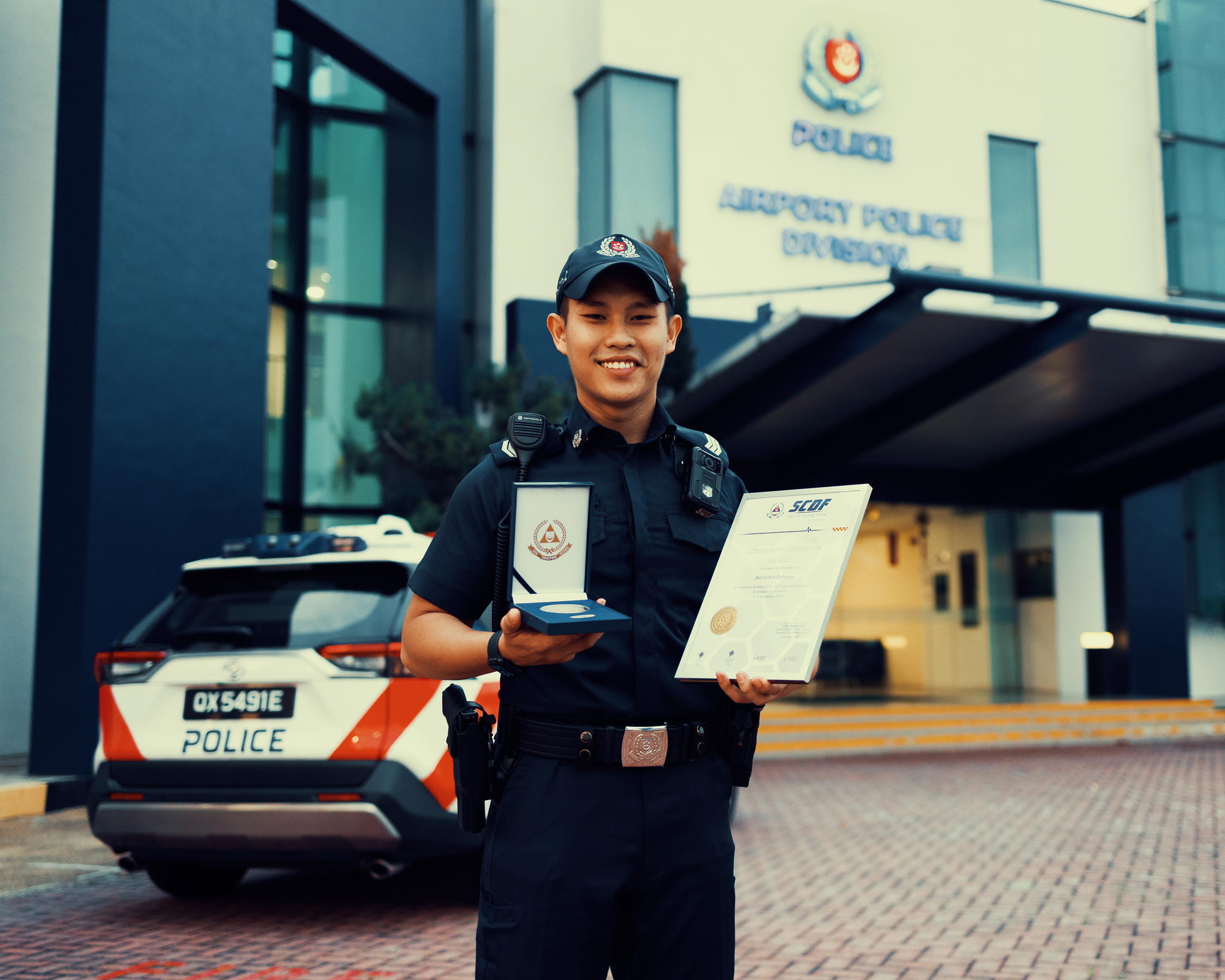 the officer is in uniform and standing infront of the airport police division, holding the certificate and the SCDF medal