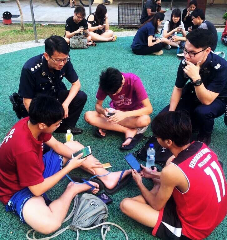 police officers interacting with a bunch of students, in their home clothes, sitting on the asphalt of the playground with handphones in the hands
