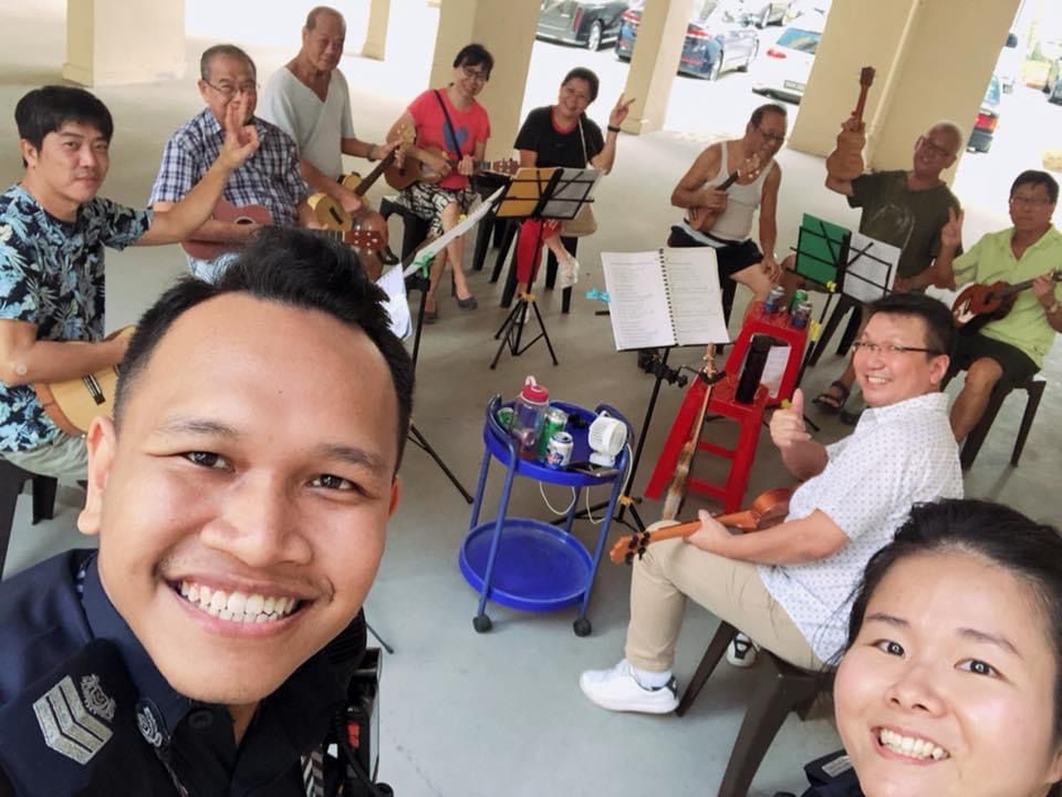 sergeant sally and a malay colleague, talking a high point selfie, with a group of elderly holding musical equipment sitting in a circle and smiling fo the camera
