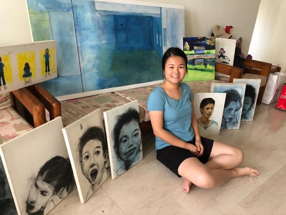 sergeant sally, in a blue t-shirt and black shorts, sitting on the floor, with canvas paintings aligned beside her