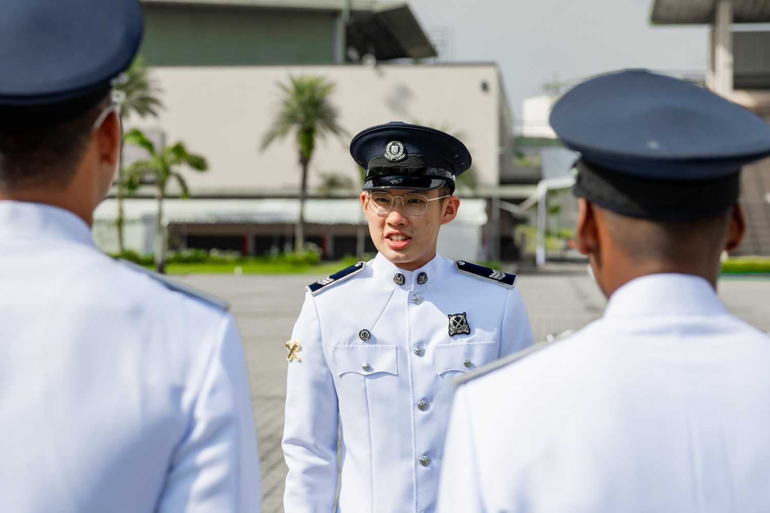 NDP Trainer Colin in white uniform and peak cap facing front and in background, talking to two officers in the foreground whose back is facing the reader