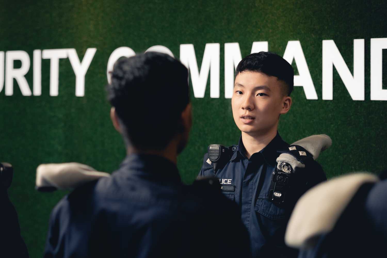 officer briefing team, facing the reader and talking to someone whose back can be seen. Behind him is a green moss background with the words "Public Transport Security Command" in white