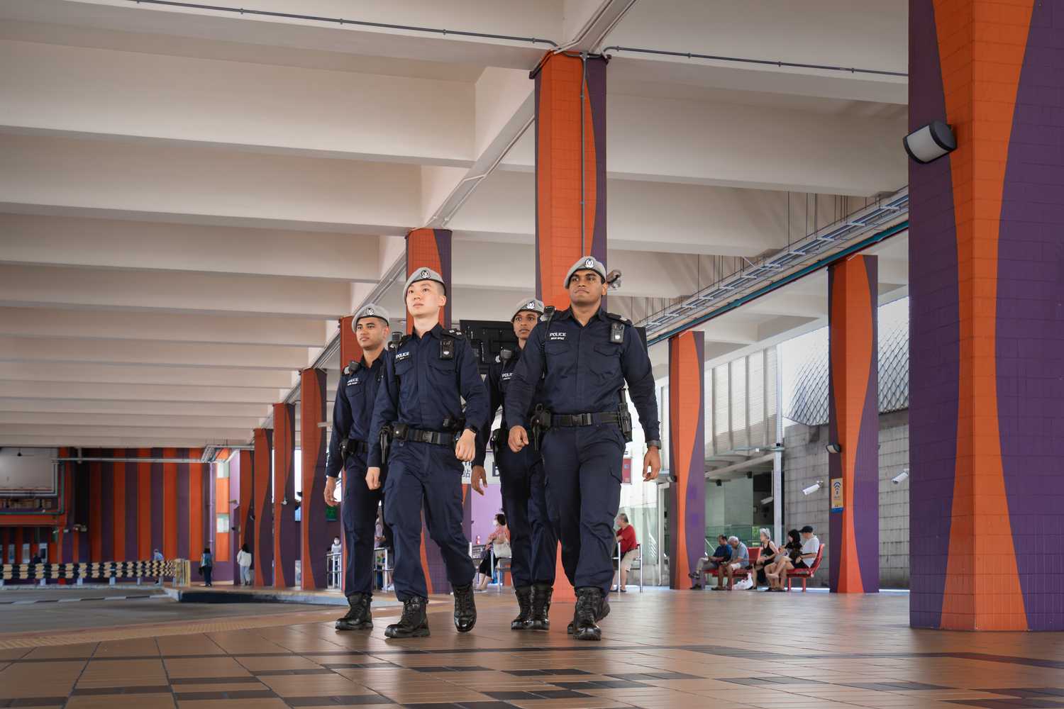 four officers walking together to the 7pm direction, at a bus interchange. The interchange is visibly orange painted with high ceilings and human traffic around
