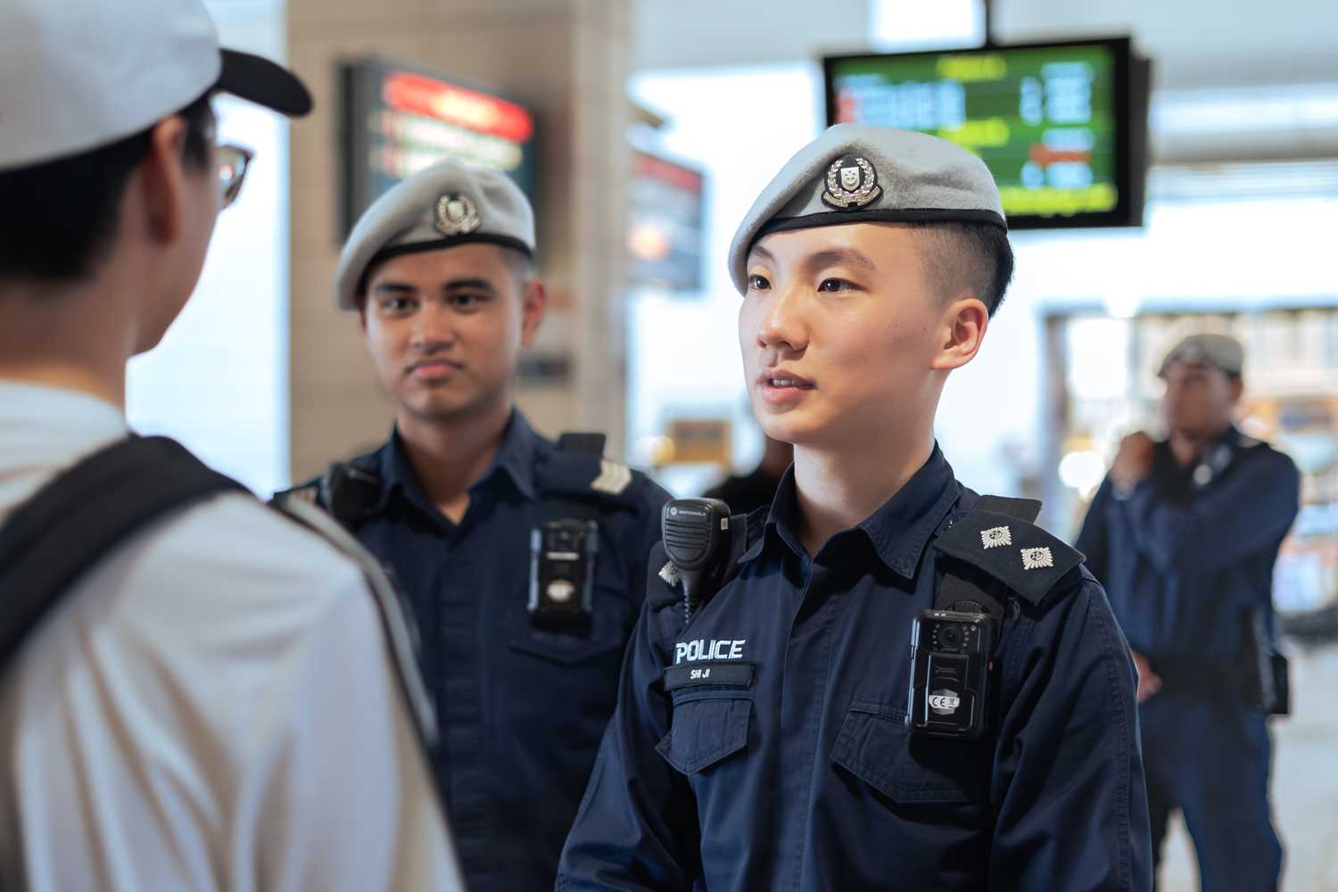 officer shi ji talking to a member of the public, who is chinese, wearing white, facing the right and carrying a black sling bag and wearing a cap