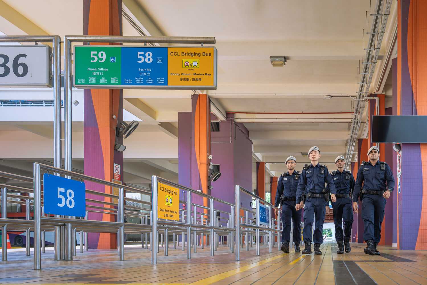 officers walking at the bus interchange but the bus signages for the waiting area can be seen, visibly in orange blue and green signboards, against an orange painted interchange with brick colour tones. 